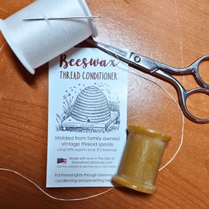 A pair of scissors, a spool of thread and hand molded beeswax thread conditioner are pictured lying on a table.