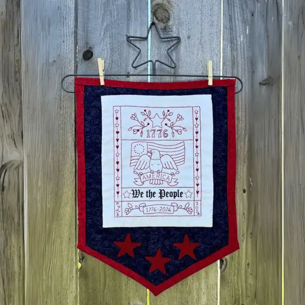 A completed and framed USA 250th Anniversary Sampler by the Posy Collection displayed on a wall.