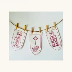 A completed trio of the Holy Family redwork embroidery kit by the Posy Collection is displayed hanging by close pins on a string.